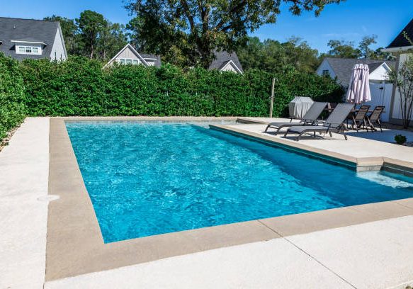 pool A rectangular new swimming pool with tan concrete edges in the fenced backyard of a new construction house with privacy hedges.